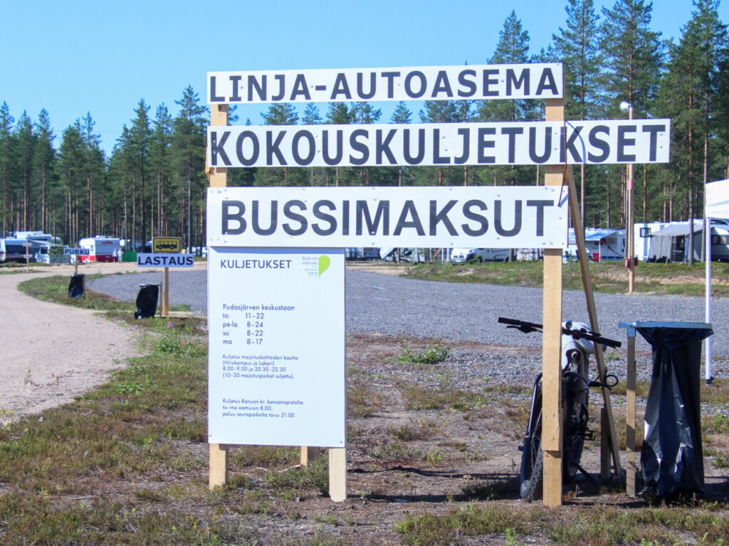 Kylttejä bussiasemalta, tolppaa vasten nojaa pyörä ja taustalla näkyy pysäköityjä asuntovaunuja.