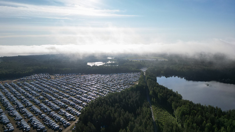 Utuinen taivaanranta, jossa paistaa aurinko. Ilmakuvassa näkyy suuri alue, jossa on matkailuautoja ja -vaunuja, metsää ja vesistöä.