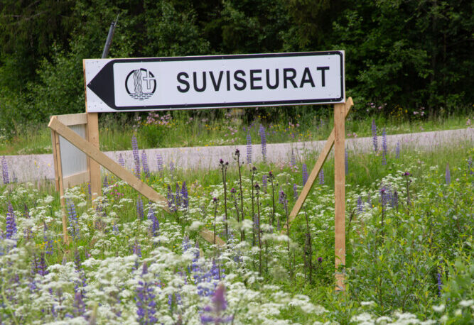 Suviseurat-opastekyltti tien reunassa, jossa kasvaa valkoisia ja violetteja kukkia.